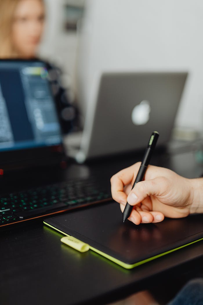Close-up of a hand using a pen tablet alongside laptops in a modern office setting.